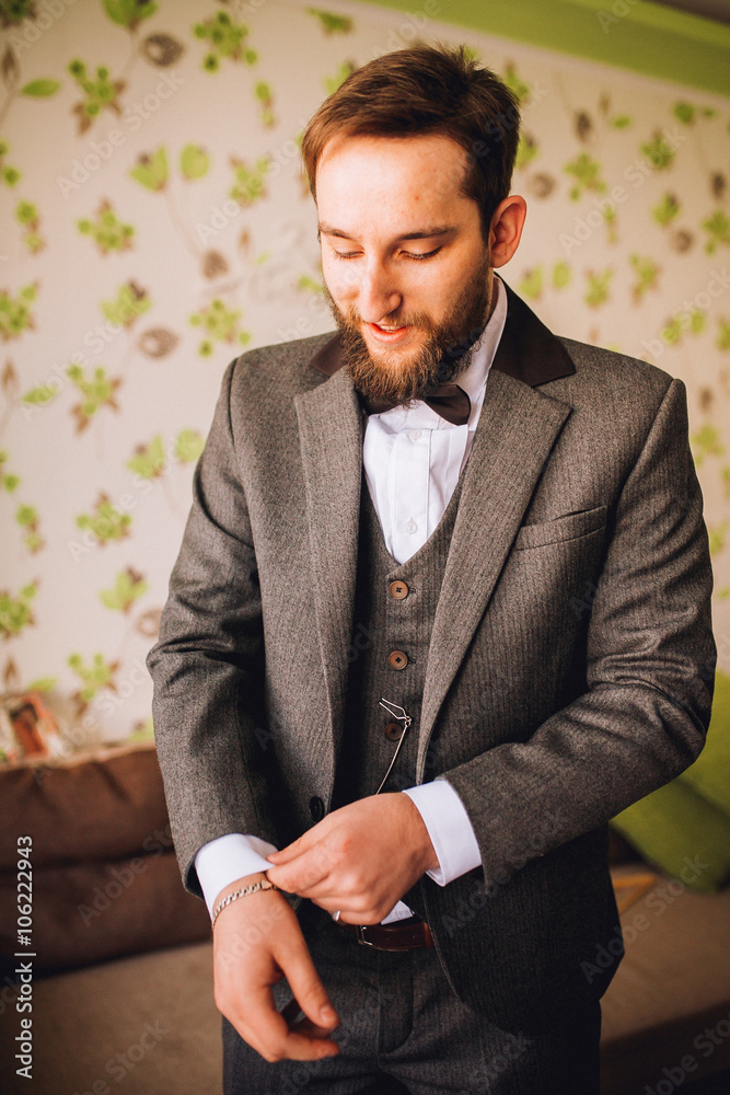 Stylish and handsome groom in waistcoat tying his bow-tie in the morning of the wedding day. Groom with beard