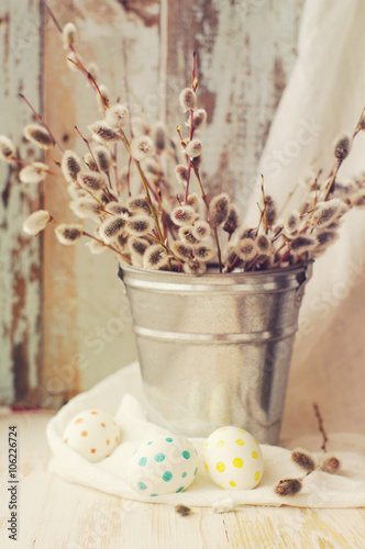 Easter eggs with a bouquet of spring branches of a willow in a bucket on a wooden background.