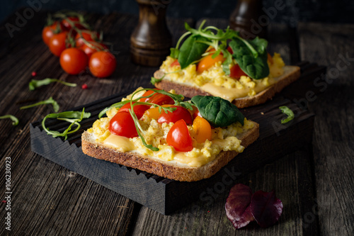 Breakfast toast with scrambled eggs, cheese, cherry tomatoes, arugula and corn salad on rustic wooden background
