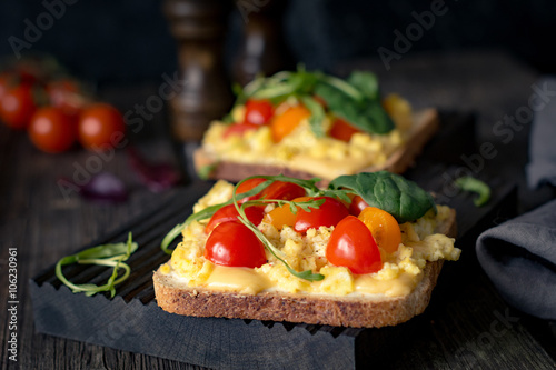 Breakfast toast with scrambled eggs, cheese, cherry tomatoes, arugula and corn salad on rustic wooden background