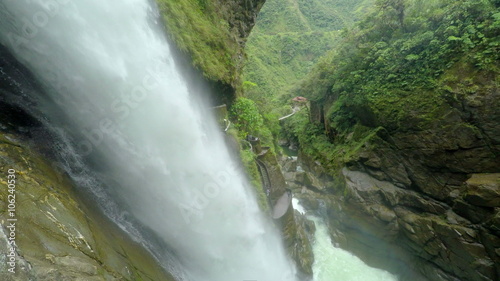 A view of the Pailon del Diablo waterfall plunging down a moss-covered cliff into a turbulent river near Banos, Ecuador, surrounded by dense green jungle and rugged rock formations, high angle.