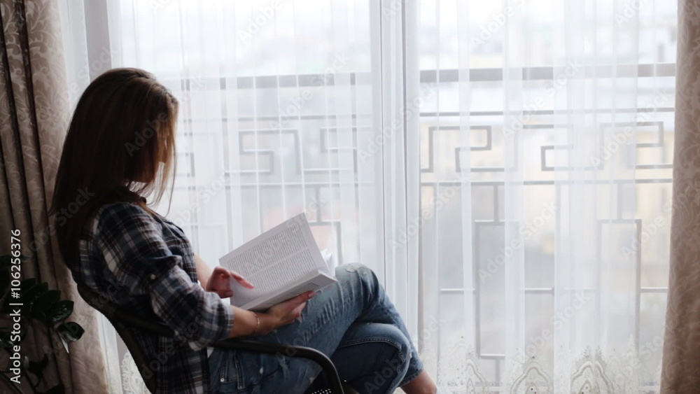 Cute woman wearing blue jeans and grey t-shirt sitting on the table in front of window and reading a book in her living room. Cute woman wearing blue jeans and grey t-shirt sitting on the table in front of window and reading a book in her living room.