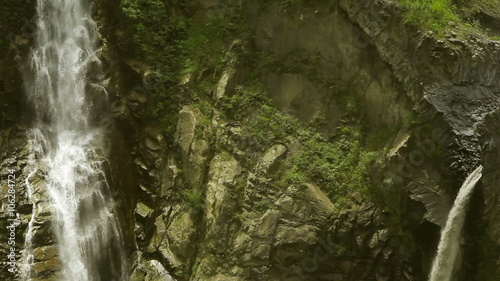 A tracking shot moves down a steep rocky mountain gorge revealing multiple waterfalls plunging over moss-covered cliffs into a pool - the Agoyan falls near Banos in the Ecuadorian Andes.