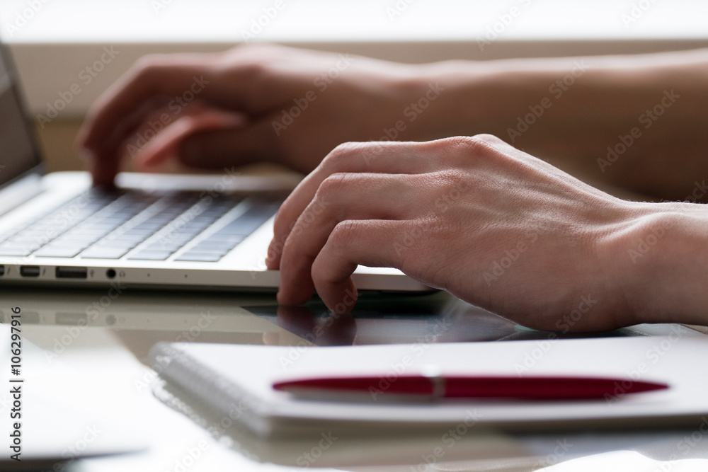 hands of a young man typing on laptop computer with notepad and pen on glass deck