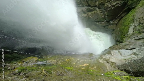 A view from within a rocky gorge as the Pailon del Diablo waterfall plunges down near Banos, Ecuador, dense mist filling the air amid vibrant green moss-covered rocks.