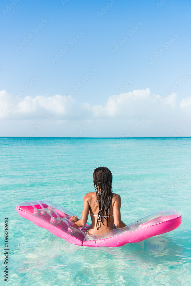 Beach vacation woman relaxing on ocean water bed. Beautiful woman from the back sitting on a pink pool float air mattress looking at view of the perfect turquoise pristine sea in tropical destination.
