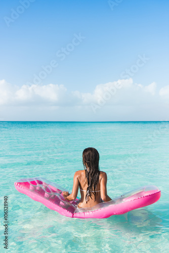 Beach vacation woman relaxing on ocean water bed. Beautiful woman from the back sitting on a pink pool float air mattress looking at view of the perfect turquoise pristine sea in tropical destination. © Maridav