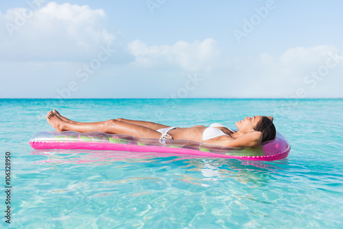 Beach sexy woman relaxing during suntan sunbathing on floating pink pool inflatable plastic air mattress float in pristine turquoise ocean water background at luxury destination getaway.