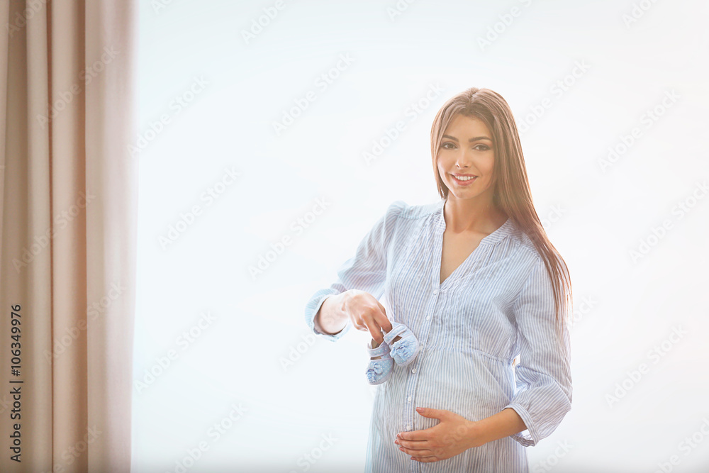 Pregnant woman standing near window with baby's bootees in the room Pregnant woman standing near window with baby's bootees in the room