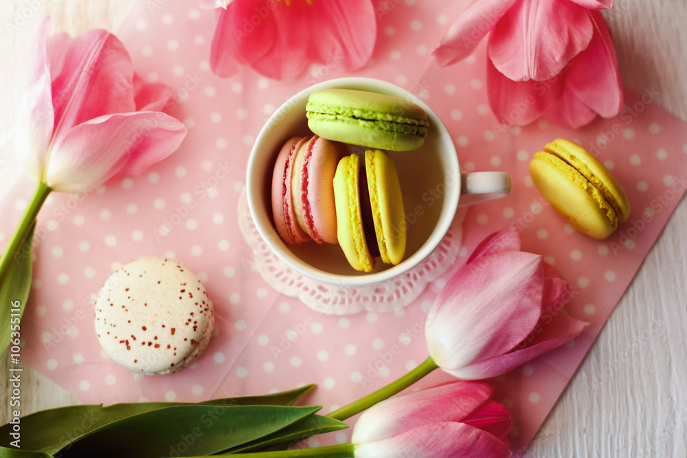 A beautiful flowers pink tulips with colorful macaroons laid in cup on white wooden background with pink Lacy napkin A beautiful flowers pink tulips with colorful macaroons laid in cup on white wooden background with pink Lacy napkin