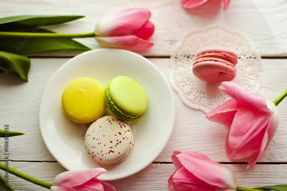 A beautiful flowers pink tulips with colorful macaroons laid on a white platter on white wooden background A beautiful flowers pink tulips with colorful macaroons laid on a white platter on white wooden background
