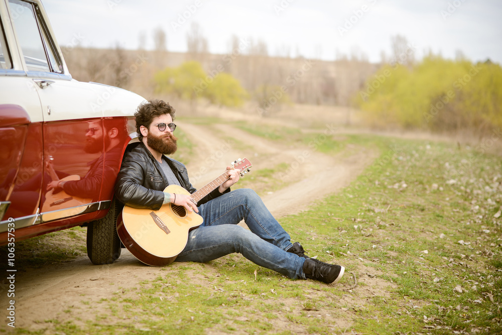 Obraz premium Bearded man playing guitar outdoors near retro car