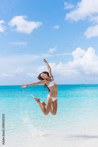 Happy bikini woman having fun jumping of joy and happiness on beach splashing water in perfect turquoise water. Asian girl cheering winning for Caribbean vacation destination during summer travel.