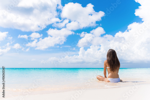 Beach vacation dream woman enjoying summer holiday on dreamy perfect ocean tropical destination. Person sitting from the back alone on deserted white sand beach getaway.