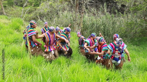 A group of indigenous men in vibrant traditional clothing perform a synchronised circle dance in tall green grass on a rocky hillside, an Inti Raymi celebration in the Ecuadorian Andes.