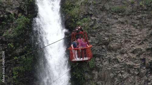 Three tourists ride a red open-air cable car across a rocky cliffside past a powerful waterfall near Banos, Ecuador, crossing the gorge framed by lush greenery and rugged rock.