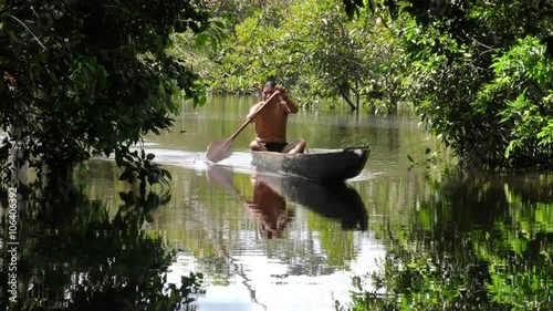 A shirtless indigenous man paddles a traditional wooden canoe along a calm reflective river in dense tropical jungle in the Cuyabeno Amazon, Ecuador, framed by overhanging trees.