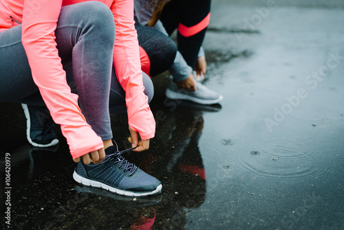 Urban athletes lacing sport footwear for running over asphalt under the rain. Two women getting ready for outdoor training and fitness exercising on cold winter weather.