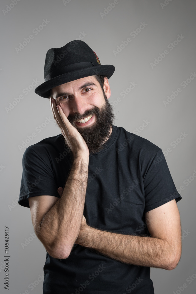Friendly smiling bearded hipster wearing black t-shirt and hat with head resting on his hand.  Headshot portrait over gray studio background with vignette. 