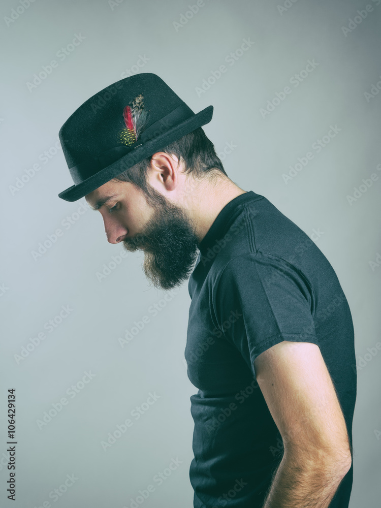 Side view of sad hunched bearded man with hat looking down. Retro toned filtered portrait over gray background with vignette effect. 