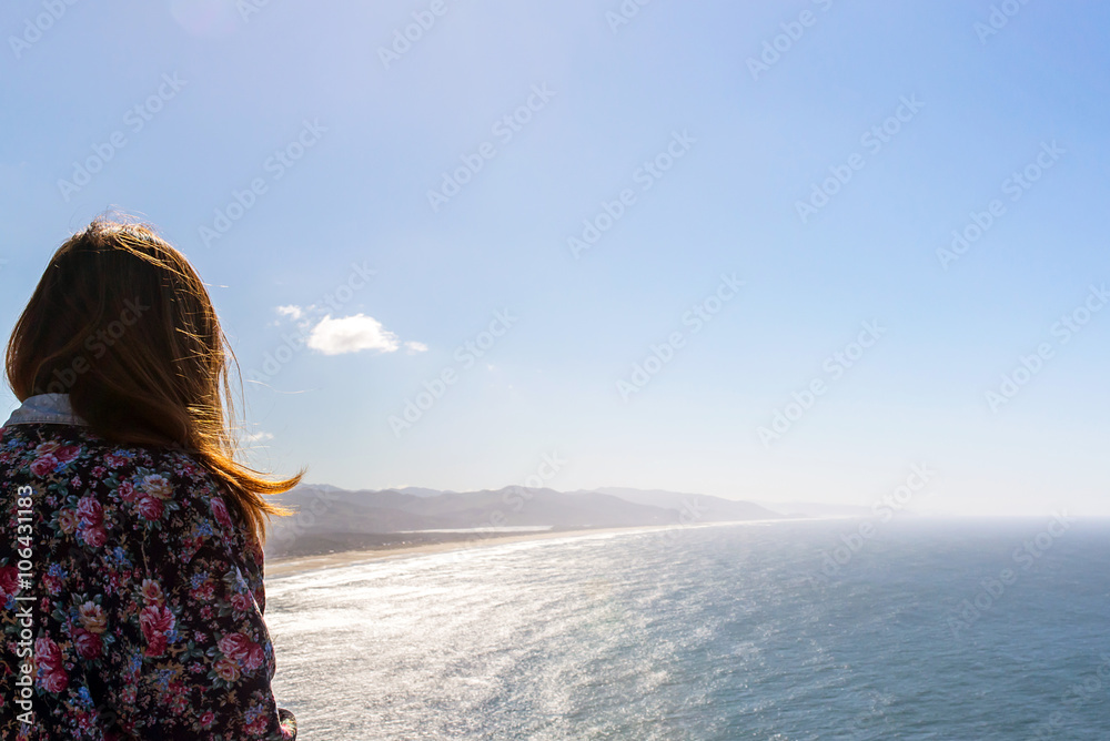 girl seen from the back with long hair and a hipster flowery coat looking at the ocean and the west coast of the united states of america during a sunny day
