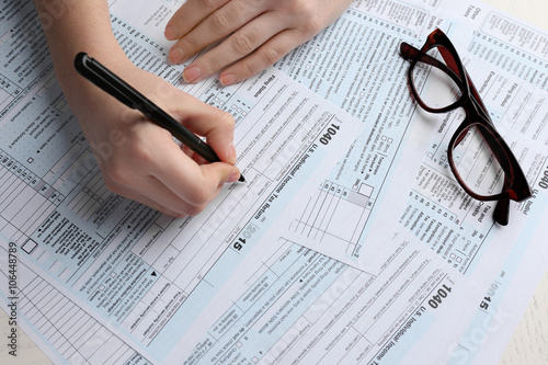 Female hand holding a pen next to the black rimmed glasses and filling in the 1040 Individual Income Tax Return Form for 2015 year on the white desk, close up photo