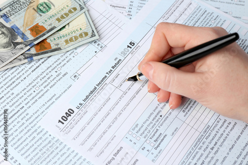Female hand holding a pen next to the metal rimmed glasses and dollar bills while filling in the 1040 Individual Income Tax Return Form for 2015 year on the white wooden desk, close up