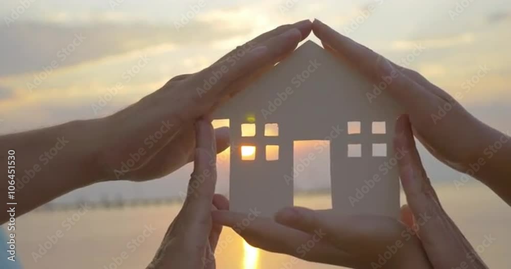Close-up shot of woman holding home model and then family making house around it with hands. Sunset and sea as background. Zooming in to get the sun in the window
