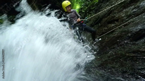 A canyoneer in a yellow helmet and wetsuit descends a powerful waterfall using ropes in lush green jungle near Banos, Ecuador, a close dynamic shot of splashing water and rugged terrain.