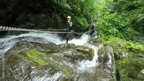 A person in helmet and harness crosses a rope bridge over a rushing river in dense green jungle near Banos, Ecuador, moss-covered rocks and cascading water around the wild scene.
