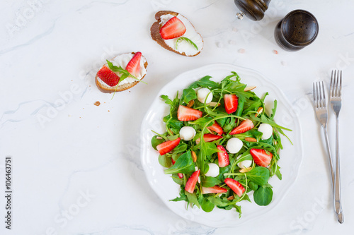 Healthy green salad with arugula, baby spinach, strawberries, almonds and mini mozzarella balls on white plate on white background, view from above