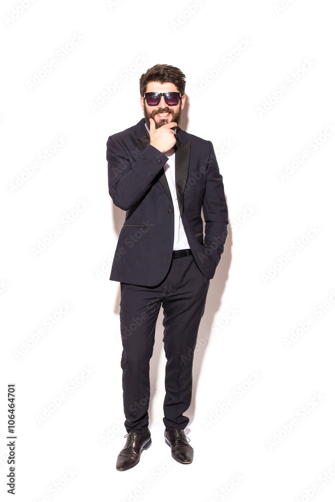 Obraz premium Full height Cheerful young handsome man in sunglasses keeping hand on chin and looking at camera with smile while standing against white background