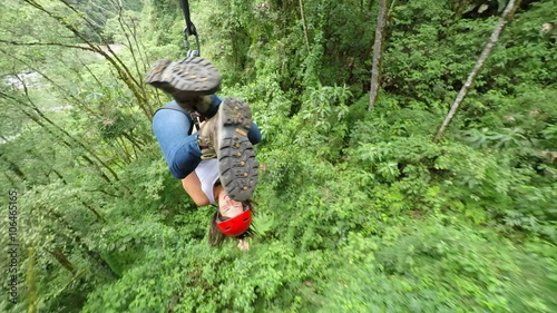 A woman in a red helmet ziplines through dense tropical jungle near Banos, Ecuador, smiling as she glides over green foliage and a rocky riverbed in a high-angle tracking shot.