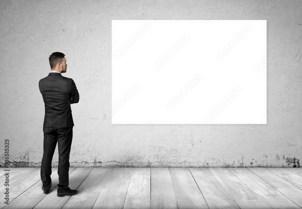 Young man in business suit, standing front of concrete wall with white blank board on it. Young man in business suit, standing front of concrete wall with white blank board on it.