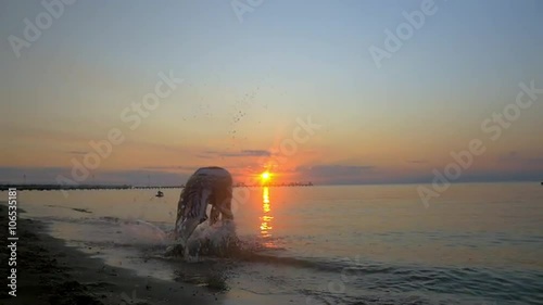 Slow motion steadicam shot of a young sportsman performing acrobatics at the seaside at sunset. He doing somersaults and falling into the water at the end photo