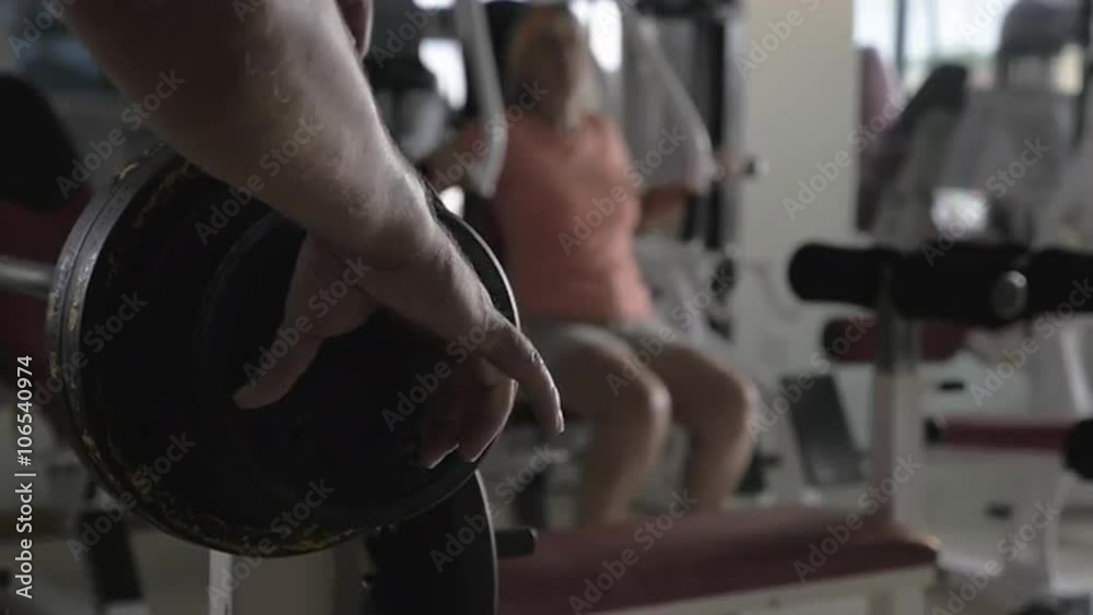 Slow motion close-up shot of fixing weight plate on the barbell, woman training on exerciser in background. Workout in fitness center