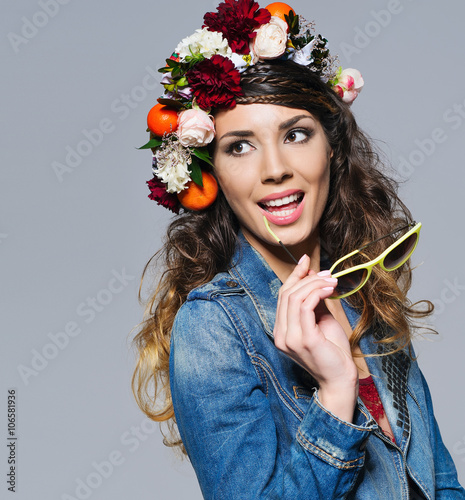 Fashion portrait of a beautiful young woman with gipsy hairstyle wearing bright flower crown, jeans jacket and trendy yellow sunglasses smiling happily