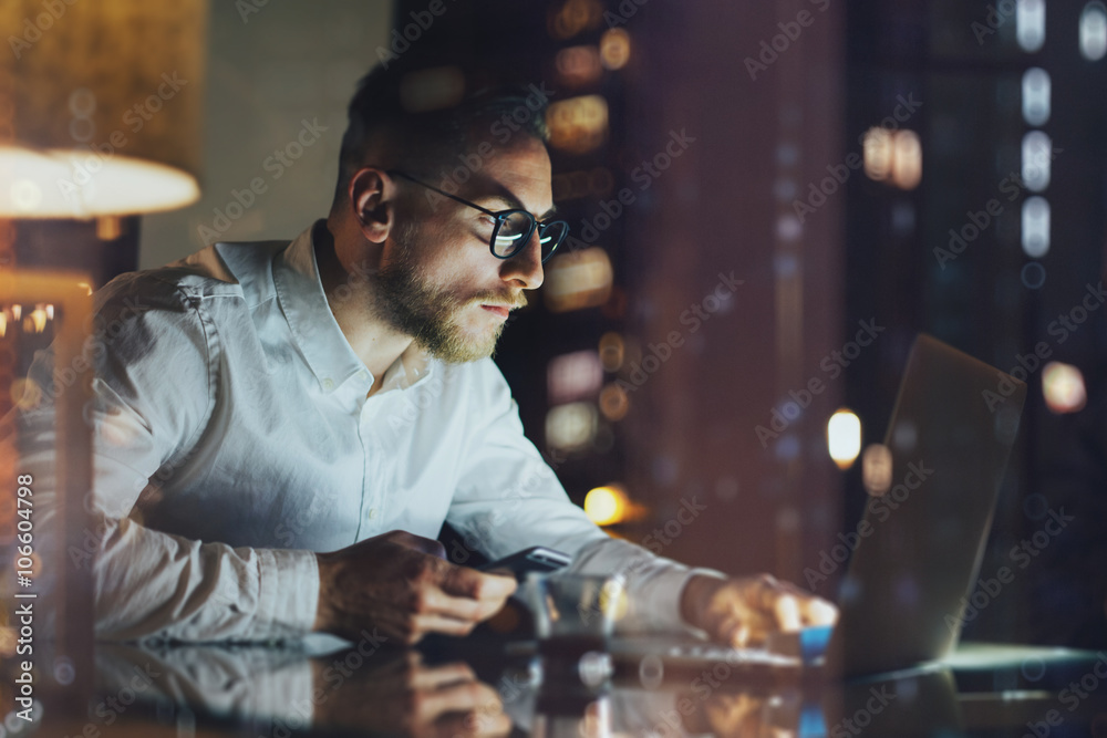 Bearded young businessman working on modern loft office at night. Man using contemporary notebook texting message, holding smartphone, bokeh background. Horizontal, film effect. Bearded young businessman working on modern loft office at night. Man using contemporary notebook texting message, holding smartphone, bokeh background. Horizontal, film effect.
