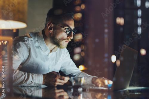 Bearded young businessman working on modern loft office at night. Man using contemporary notebook texting message  holding smartphone  bokeh background. Horizontal  film effect. 