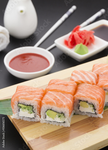 Salmon sushi roll on wooden plate ( gete ) with ginger wasabi chopsticks and sauces over black background