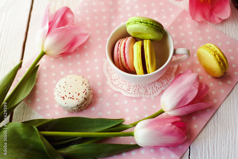 A beautiful flowers pink tulips with colorful macaroons laid in cup on white wooden background with pink Lacy napkin A beautiful flowers pink tulips with colorful macaroons laid in cup on white wooden background with pink Lacy napkin