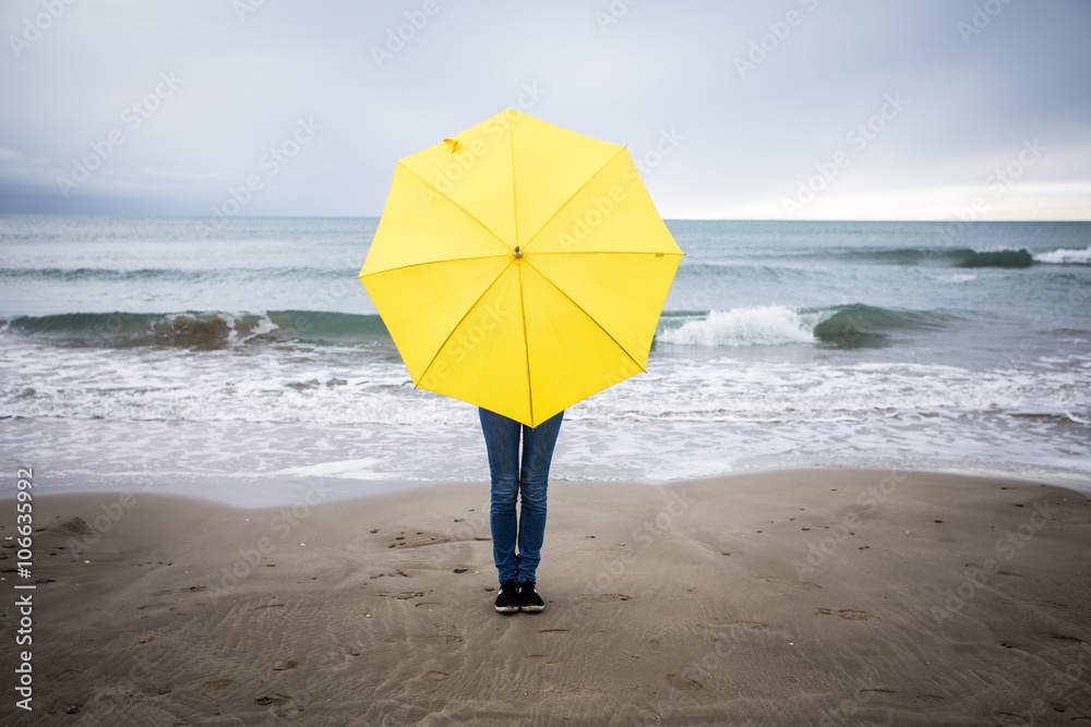 Une personne sur une plage caché derrière un parapluie jaune soleil au centre de l'image