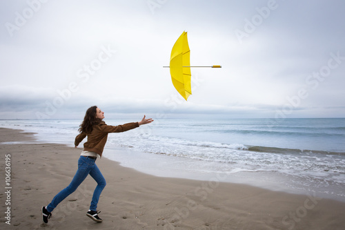 Un jeune  femme tentant de rattraper son parapluie jaune envolé sur une plage  photo