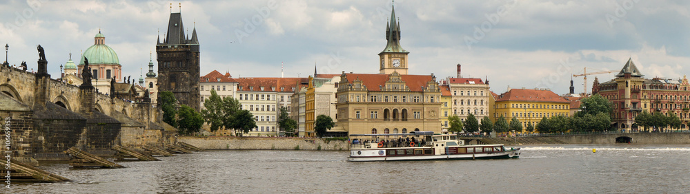 Wide panorama looking towards the eastern bank of Prague’s Vlatava river. Charles Bridge, Saint Francis of Assisi Church, Old Town Bridge Tower, an historic water tower and a river cruise boat.