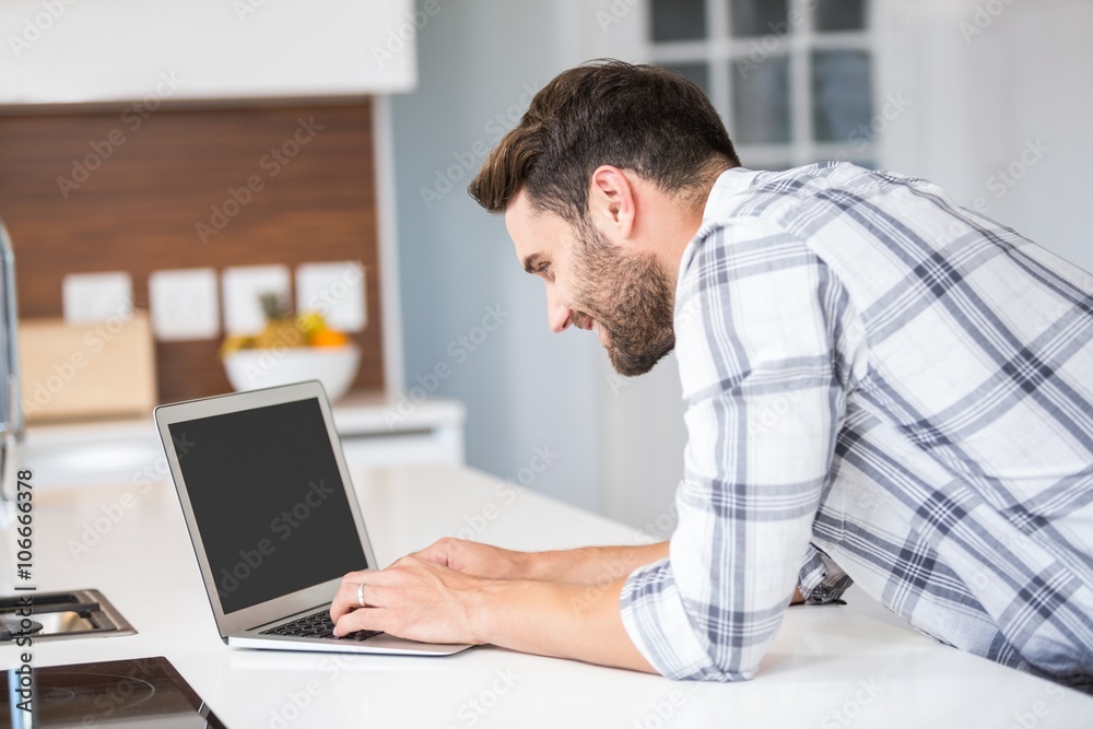 Happy young man using laptop while leaning on kitchen counter