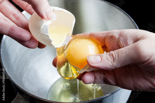 Separating yolk and white of the white shell egg in woman hand above metal stainless steel mixing bowl on black background