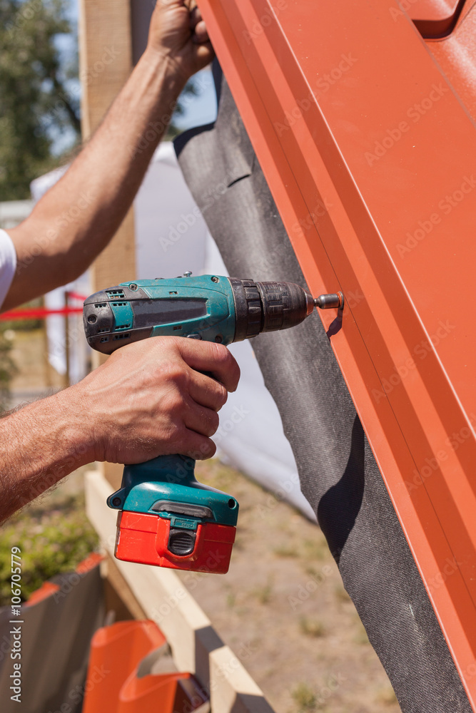 Worker on a roof with electric drill installing red metal tile on wooden house Worker on a roof with electric drill installing red metal tile on wooden house