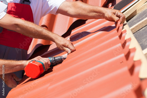 Workers on a roof with electric drill installing red metal tile on wooden house