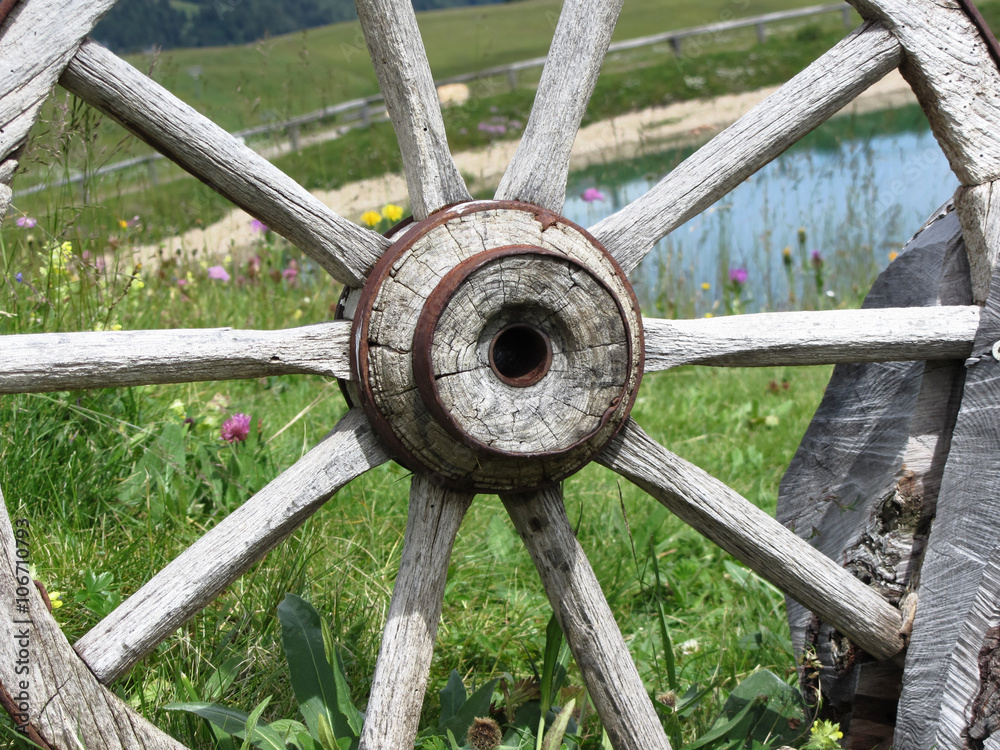 Old wooden wagon wheel with alpine lake and pasture in background Old wooden wagon wheel with alpine lake and pasture in background