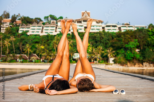Two beautiful girls in swimsuits lying on the wooden pier upside down, against the background of a hotel on the beach photo